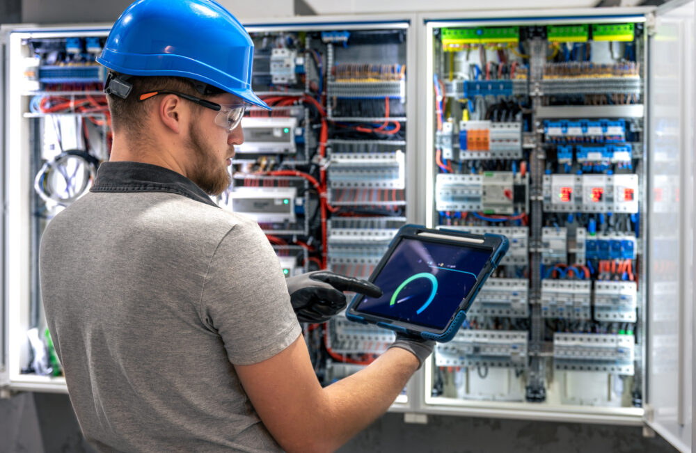 A technician in safety gear stands with a tablet in front of an open switchboard, analyzing data. Industrial digital monitoring scene.